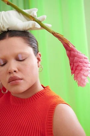 Portrait of an adult female with closed eyes, wearing an orange sleeveless top, posed with a pink flower and white gloves against green.