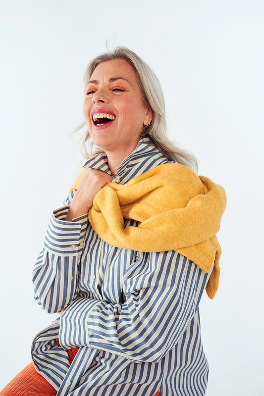 Cheerful woman with silver hair and striped shirt laughing against a white backdrop, her yellow scarf casually draped over her shoulders.