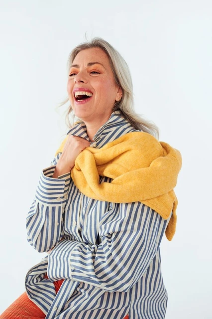 Cheerful woman with silver hair and striped shirt laughing against a white backdrop, her yellow scarf casually draped over her shoulders.