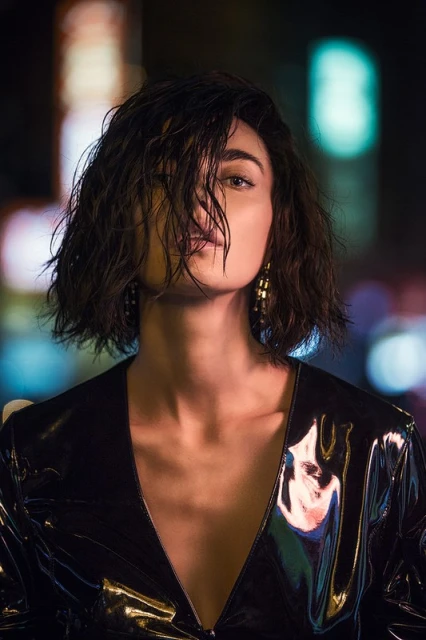 Close-up portrait of a brunette adult woman with short, wet hair wearing earrings and a shiny, black v-neck top against a blurred background.