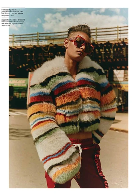 Male fashion model wearing a colorful striped faux fur jacket and red pants poses on a city street under an elevated train track.
