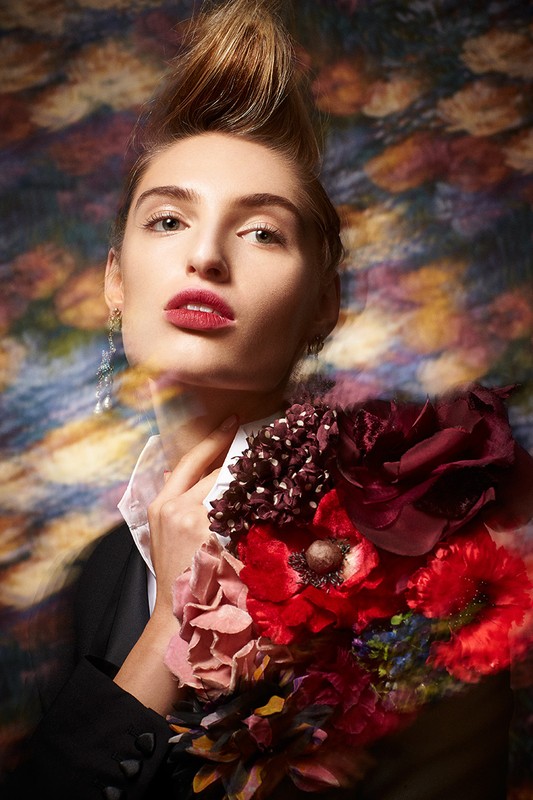 Elegant woman with stylized blonde hair and red lipstick holds a bouquet of red flowers against a blurred abstract background.