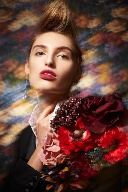 Elegant woman with stylized blonde hair and red lipstick holds a bouquet of red flowers against a blurred abstract background.