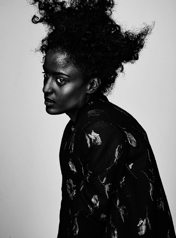 Monochrome portrait of a Black woman with curly afro hair, wearing a patterned jacket, shot in a photo studio with soft lighting.