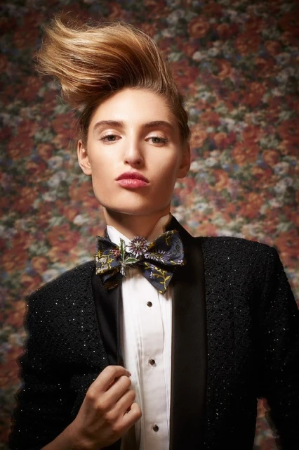 Elegant woman in tuxedo-style jacket and statement bow tie against a floral backdrop, styled with a high-volume hairstyle and bold expression.