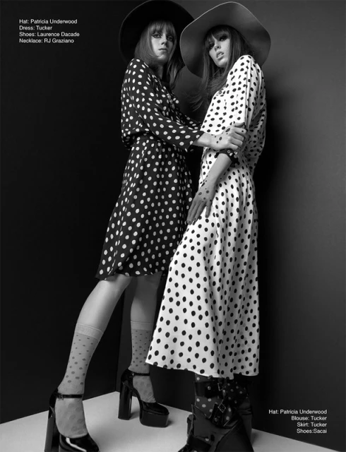 Two fashion models pose in polka dot dresses and hats against a dark background in a studio fashion shot. Monochrome styling.