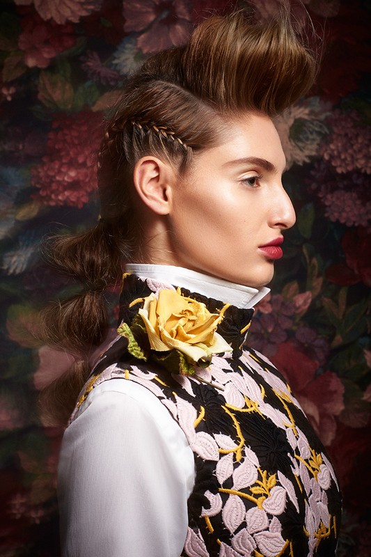 A woman with sculpted brown hair and a yellow rose on her collar poses in a close-up studio shot against a floral background.