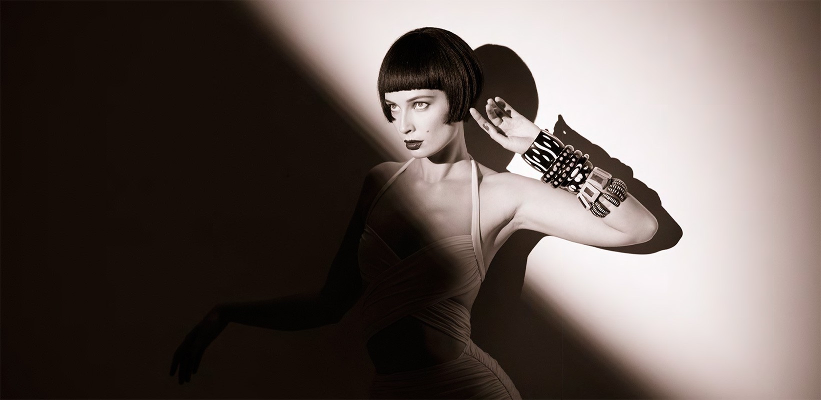 An adult woman with a bob haircut poses indoors. She is wearing a dress and multiple bracelets and is lit with dramatic lighting.