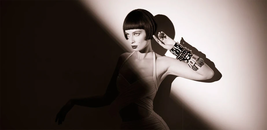 An adult woman with a bob haircut poses indoors. She is wearing a dress and multiple bracelets and is lit with dramatic lighting.