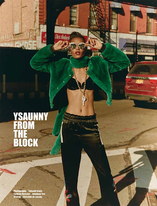 Fashionable woman with sunglasses, green jacket, and gold jewelry, posing on a city street with urban buildings and a parked car.