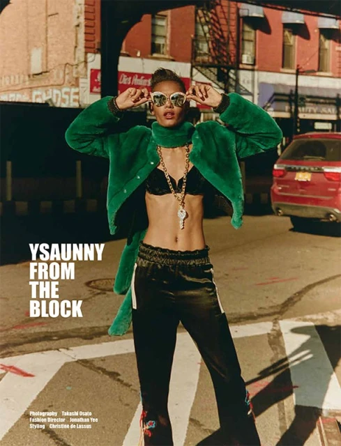 Fashionable woman with sunglasses, green jacket, and gold jewelry, posing on a city street with urban buildings and a parked car.