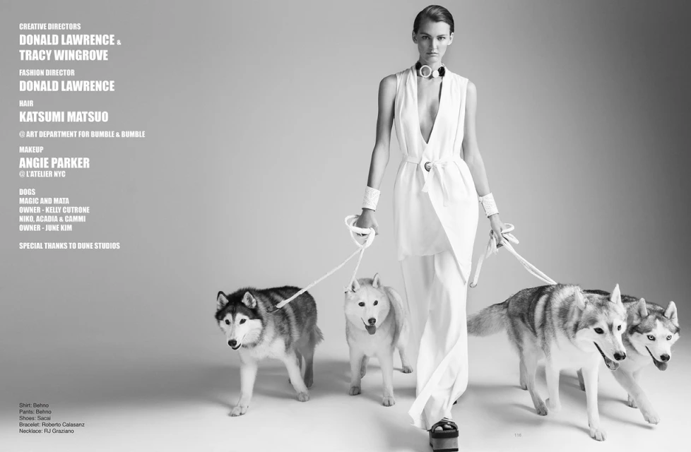 Monochrome fashion shot of a model in white walking four dogs on leashes against a gray backdrop, showcasing fashion and animal companionship.