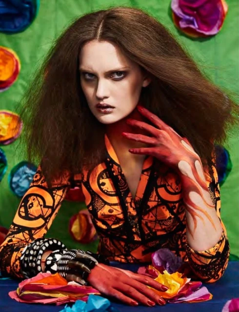 A model with painted arms poses in front of a green backdrop with colorful decorative circles, wearing a patterned jacket and dark lipstick.