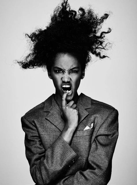 Striking black and white portrait of an adult female with voluminous afro-textured hair, wearing an oversized blazer, making an intense expression.