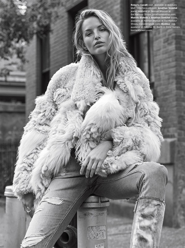 A woman in a fur coat and ripped jeans poses in an urban setting with brick buildings. The photograph is black and white, highlighting the texture of the coat.