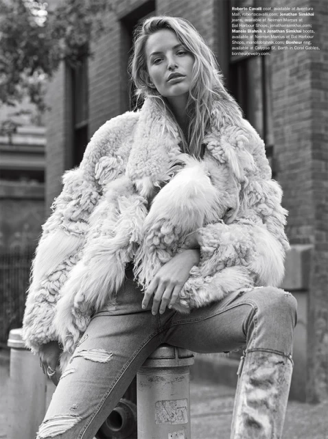A woman in a fur coat and ripped jeans poses in an urban setting with brick buildings. The photograph is black and white, highlighting the texture of the coat.