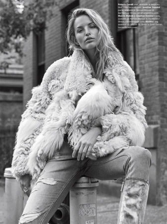 A woman in a fur coat and ripped jeans poses in an urban setting with brick buildings. The photograph is black and white, highlighting the texture of the coat.