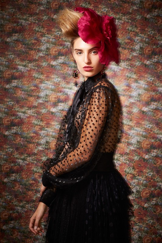Portrait of a woman with an elaborate updo and floral headpiece, wearing a sheer, polka-dot blouse and tiered skirt against a floral background.