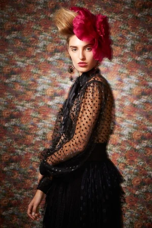 Portrait of a woman with an elaborate updo and floral headpiece, wearing a sheer, polka-dot blouse and tiered skirt against a floral background.