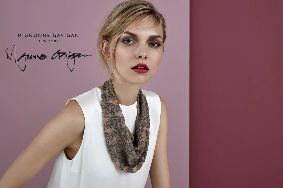 A blonde woman wears a sleeveless white dress and patterned necklace against a split pink and maroon background in a studio shot.