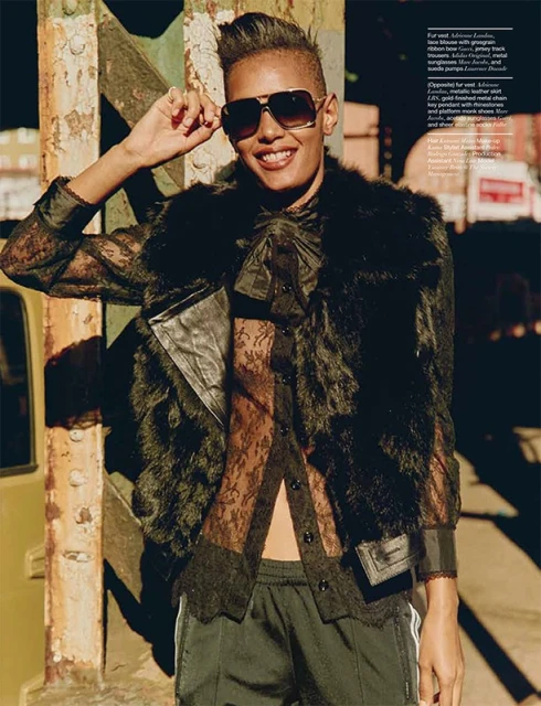 Fashionable adult with short hair, wearing sunglasses, fur coat, and athletic pants, poses outdoors against an urban backdrop.