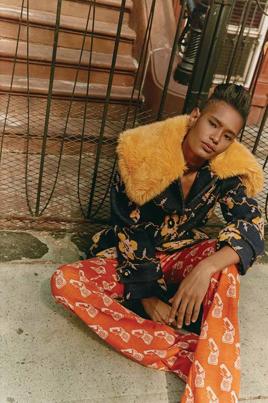 A person sitting outdoors on stone stairs with a jacket with a fur collar and patterned pants, and metal fencing in the background.