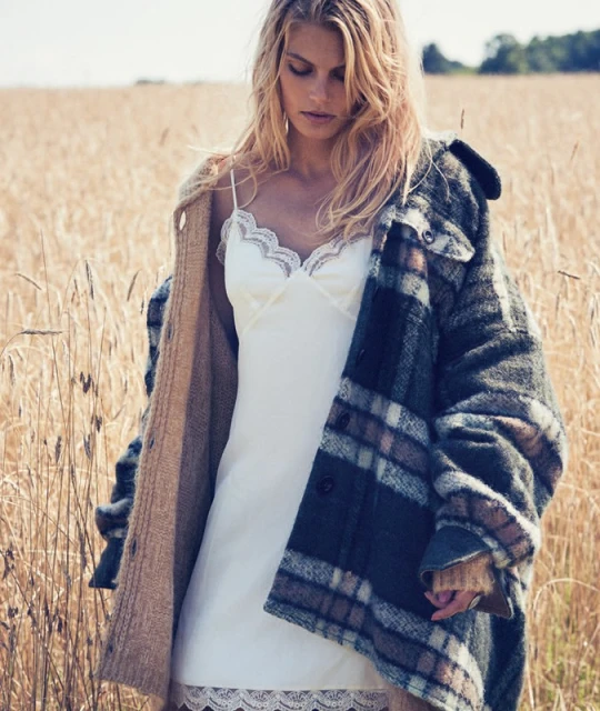 A blonde woman in a white slip dress and plaid jacket stands in a wheat field on a sunny day, creating a natural and stylish look.