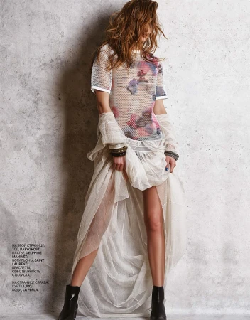 Model in mesh floral top and layered sheer white skirt with black boots standing against textured grey wall