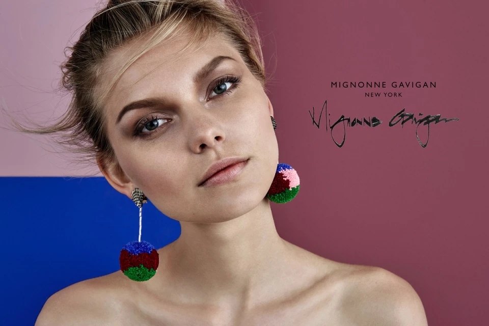 Close-up studio shot of a blonde woman wearing Mignonne Gavigan New York colorful pompom earrings, against a pink and blue background.