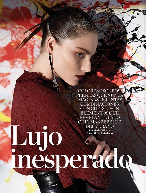 Close-up editorial fashion shot of a young adult woman with dark hair and dark eye makeup, wearing a burgundy top, against an abstract background.