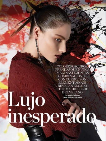 Close-up editorial fashion shot of a young adult woman with dark hair and dark eye makeup, wearing a burgundy top, against an abstract background.