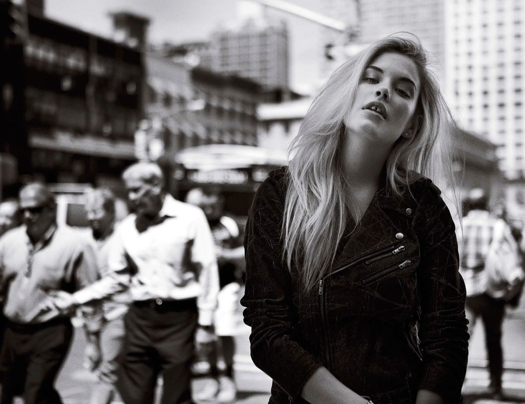 Black and white portrait of a blonde woman in a dark jacket posing outdoors on a busy city street.