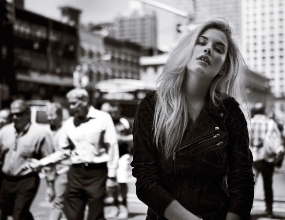Black and white portrait of a blonde woman in a dark jacket posing outdoors on a busy city street.