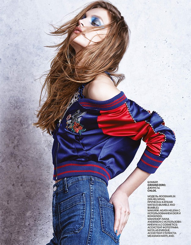 A young adult woman poses in a fashion shot, wearing a blue and red bomber jacket with jeans against a concrete background in a studio setting.