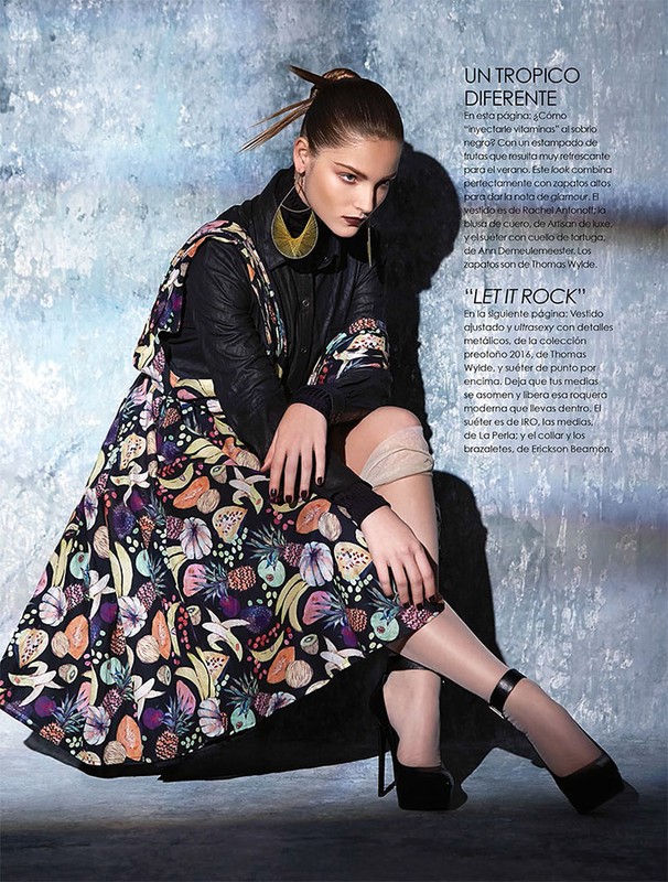 Fashion editorial image: a woman poses indoors with dark top, fruit-print skirt, large earrings, and high heels against a mottled wall.