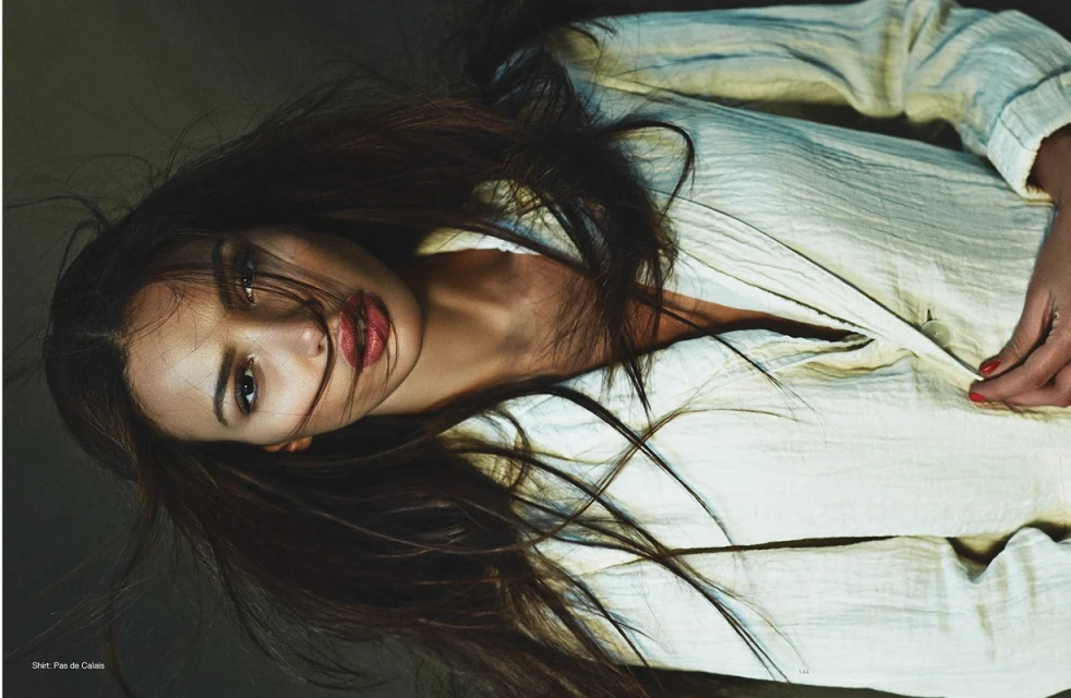Brunette woman in a white shirt looks at the camera with dark background and dark hair. Red lipstick and red nail polish detail.