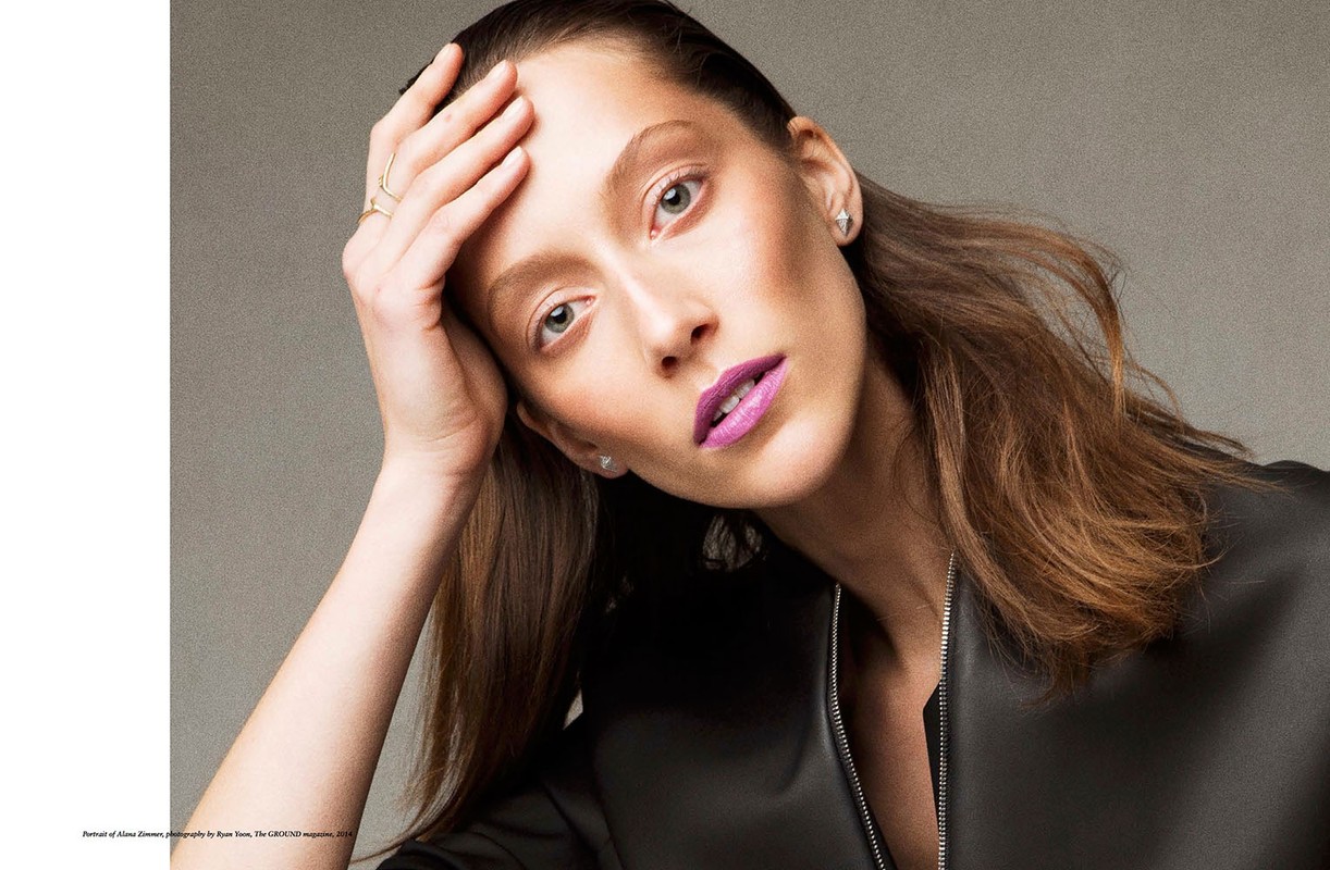 Close-up portrait of an adult woman with brown hair and lilac lipstick looking at the camera in a studio setting with soft lighting.
