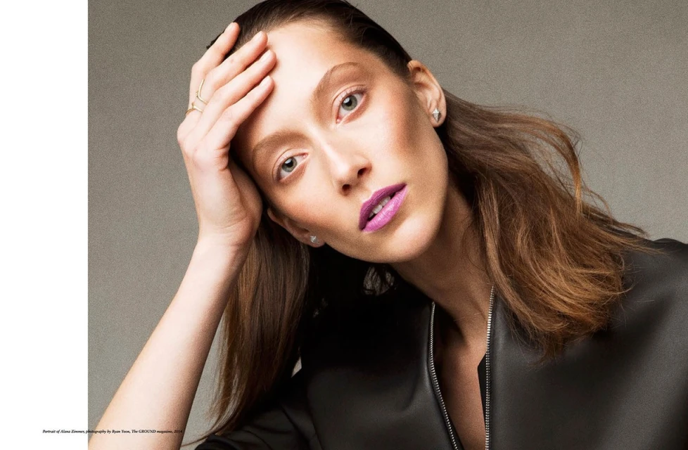 Close-up portrait of an adult woman with brown hair and lilac lipstick looking at the camera in a studio setting with soft lighting.
