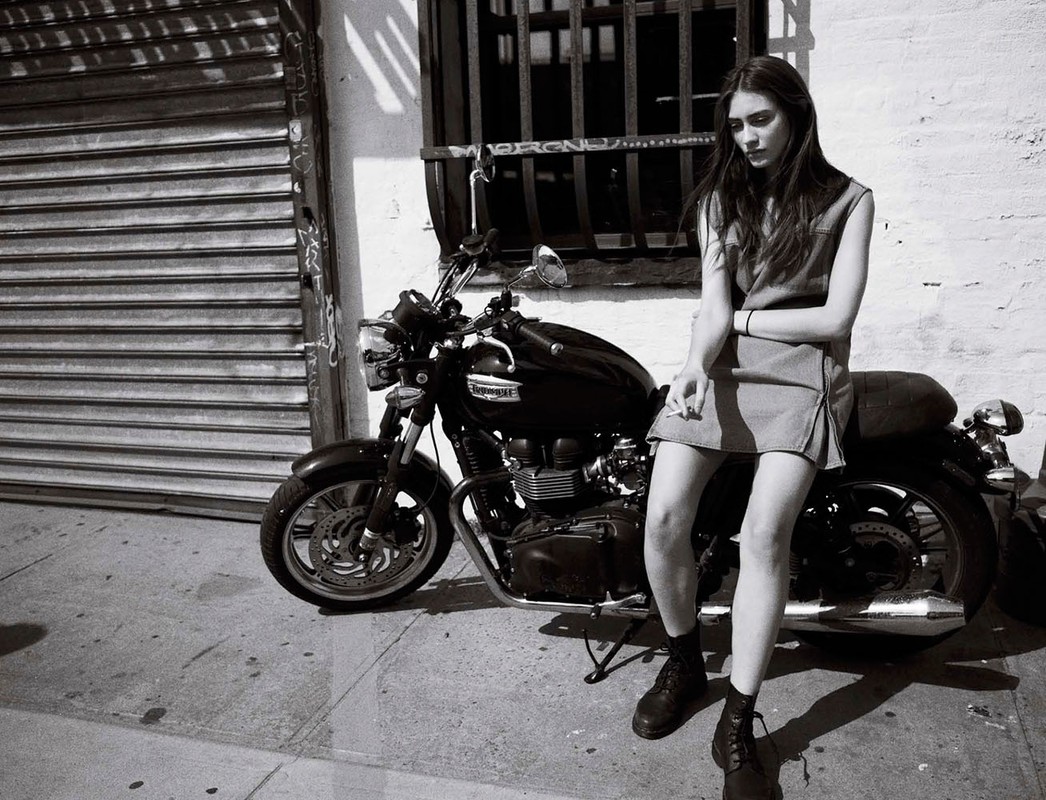 Black and white shot of a stylish woman with long dark hair sitting on a vintage motorcycle in an urban setting, posing with a cigarette.