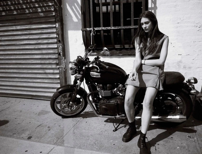 Black and white shot of a stylish woman with long dark hair sitting on a vintage motorcycle in an urban setting, posing with a cigarette.