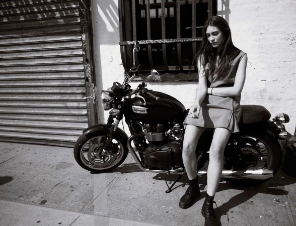 Black and white shot of a stylish woman with long dark hair sitting on a vintage motorcycle in an urban setting, posing with a cigarette.
