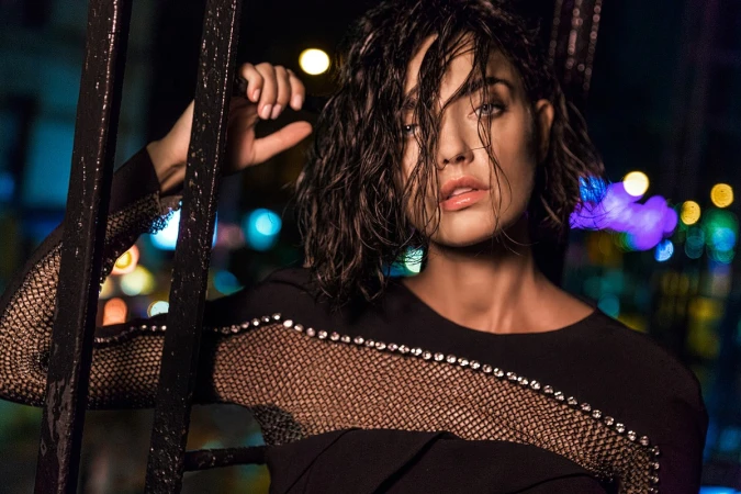 Close-up portrait of a woman with wet dark hair in a black mesh-paneled top outdoors at night with blurred colorful city lights