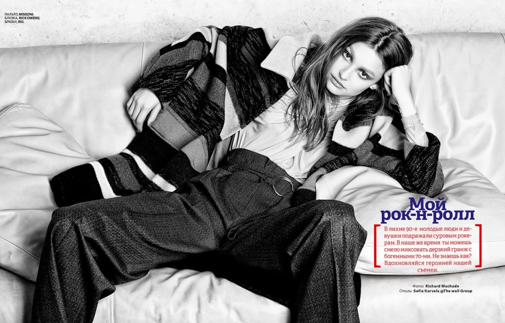 Monochrome image of a young adult female reclining on a white couch, styled in an oversized patterned jacket and dark trousers in a studio.