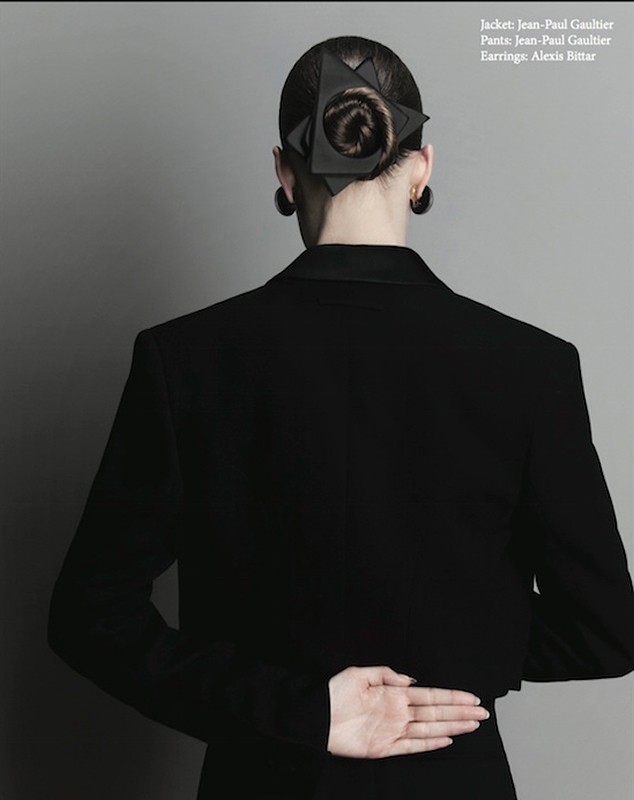 Back view of a woman with dark hair styled in a bun and secured with a geometric hair accessory, wearing a black blazer.