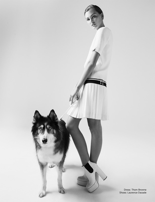 A stylish adult female poses in a studio with a dog. She wears a fashionable white outfit and platform shoes against a neutral background.