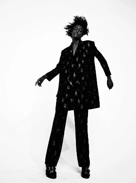 A black model poses in a patterned, oversized black blazer and matching pants against a bright white backdrop in a studio setting.