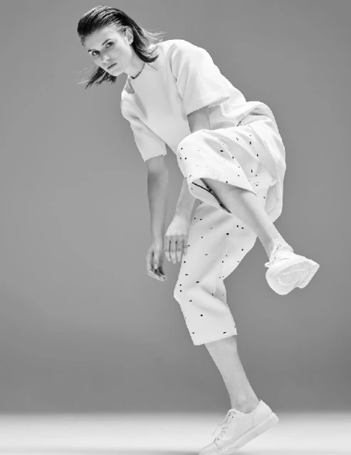 Fashion portrait of an adult woman in light clothing, posing in a dynamic stance against a grey backdrop, captured in black and white.