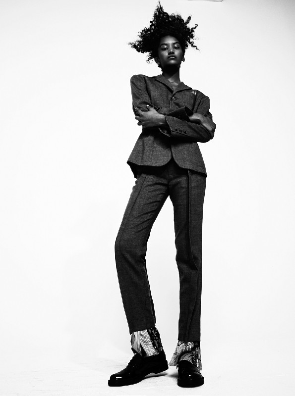 Black and white fashion shot: a woman in a grey suit stands with arms crossed, wearing black shoes in a photo studio setting.