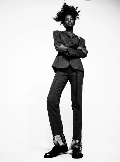 Black and white fashion shot: a woman in a grey suit stands with arms crossed, wearing black shoes in a photo studio setting.