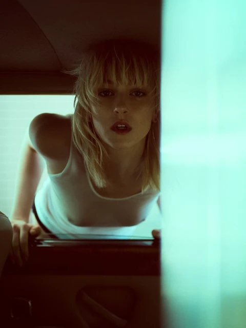 Indoor portrait of a blonde woman in a white top, posing next to a blurred turquoise vertical object inside a dark room, lit by a soft light.